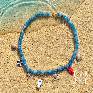 May include: A beaded necklace featuring a strand of translucent blue beads. The necklace is adorned with various charms including a white and blue life preserver, a metallic starfish, a white fish, a metallic turtle, a red lobster, and seashells.