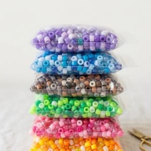 Puede incluir: Una pila vertical de seis bolsas de plástico transparente llenas de pequeñas cuentas cilíndricas de colores variados. Las bolsas contienen cuentas moradas, azules, marrones, verdes, rosas y naranjas, con acabados sólidos, translúcidos y brillantes.