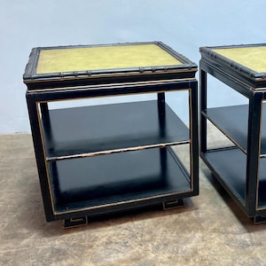 May include: Two matching black side tables with a square shape. Each table has a gold-colored top and two shelves. The tables have a bamboo-style trim and are placed side by side.