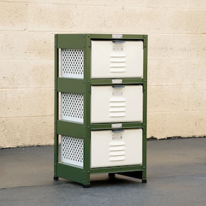Op de afbeelding: Een drielaags metalen opbergunit met een groen frame en witte metalen lades. Elke lade heeft een klein metalen handvat en een rasterpatroon aan de voorkant.
