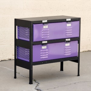 Op de afbeelding: Een metalen locker met twee verdiepingen met twee paarse lades en zwarte metalen frames. De locker heeft een zwarte metalen bovenkant.