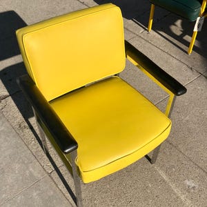Op de afbeelding: Een retro stoel met een heldergele zitting en rugleuning. De stoel heeft zwarte armleuningen en een verchroomd metalen frame. Op de achtergrond is een andere stoel met een groene zitting te zien.