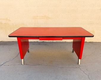 May include: A red metal desk with a black trim and a single drawer. The desk has chrome legs and is sitting on a concrete floor.