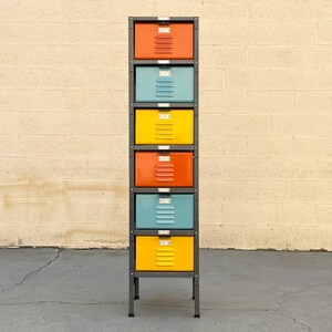 May include: A tall metal storage unit with six drawers. The drawers are painted in alternating colors of yellow, blue, and orange. The unit is standing on a gray concrete surface in front of a beige brick wall.