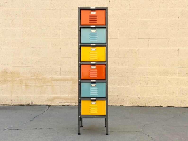 May include: A tall metal storage unit with six drawers. The drawers are painted in alternating colors of yellow, blue, and orange. The unit is standing on a gray concrete surface in front of a beige brick wall.