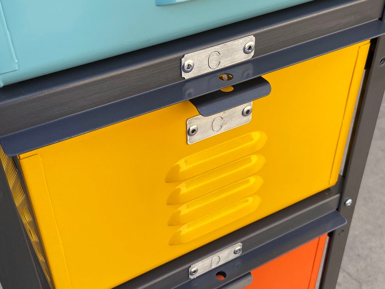 May include: A yellow metal locker with a black metal frame and a handle. The locker has a metal label with the letter "C" on it. The locker is part of a stack of lockers.