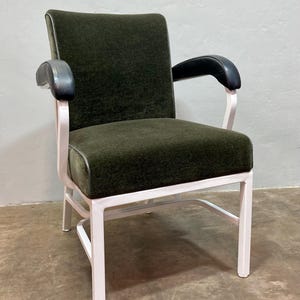 May include: An upholstered armchair with a dark green seat and back, featuring black armrests and a white metal frame. The chair has a mid-century modern design.
