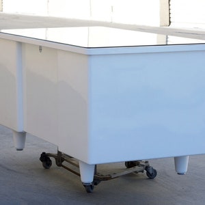 May include: White desk with a glass top and four legs. The desk is on wheels and has a small drawer on the front.