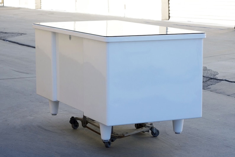 May include: White desk with a glass top and four legs. The desk is on wheels and has a small drawer on the front.