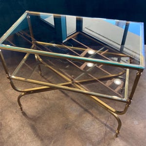 May include: A rectangular coffee table with a clear glass top and a gold-coloured metal frame. The frame has an intricate, geometric design with curved legs. The table has a modern, elegant aesthetic.
