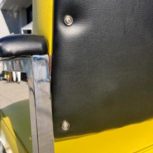 Op de afbeelding: Close-up van een gele en zwarte stoel met een chromen armleuning. De rugleuning is zwart met twee zilveren klinknagels. Het zitkussen is geel. De armleuning is zwart.