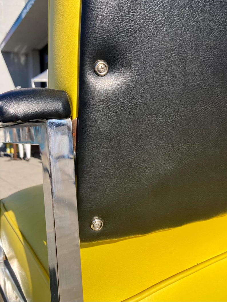 Op de afbeelding: Close-up van een gele en zwarte stoel met een chromen armleuning. De rugleuning is zwart met twee zilveren klinknagels. Het zitkussen is geel. De armleuning is zwart.