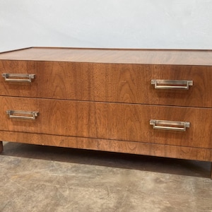 Könnte beinhalten: Zweischubladen-Kommode aus Holz mit warmbraunem Finish. Sie hat rechteckige Metallgriffe und steht auf kurzen, quadratischen Beinen. Die Holzmaserung ist sichtbar und verleiht dem Stück Textur.