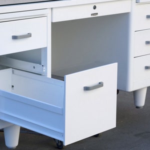 May include: White metal desk with a glass top and multiple drawers. The bottom drawer is open and the desk has metal legs.
