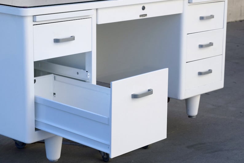 May include: White metal desk with a glass top and multiple drawers. The bottom drawer is open and the desk has metal legs.