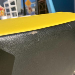 Op de afbeelding: Close-up van een geel en zwart leren object. De bovenkant is felgeel en de onderkant is zwart. Het leer heeft een getextureerde uitstraling. Er zijn een paar kleine onvolkomenheden op het zwarte oppervlak.