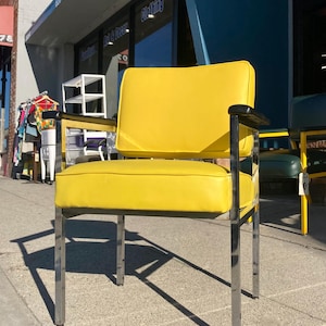 Op de afbeelding: Een retro stoel met een felgele zitting en rugleuning. De stoel heeft een chromen frame en zwarte armleuningen. De stoel staat op een stoep voor een winkel.