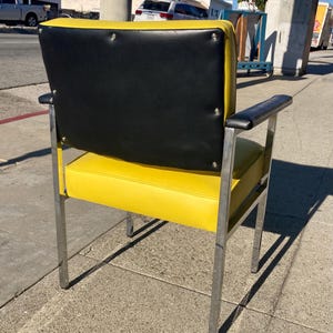 Op de afbeelding: Een retro-stoel met een heldergele zitting en rugleuning, gecontrasteerd door een zwarte rugleuning. De stoel heeft een verchroomd frame en armleuningen, wat een modern design uit het midden van de eeuw laat zien. De stoel staat buiten op een stoep.