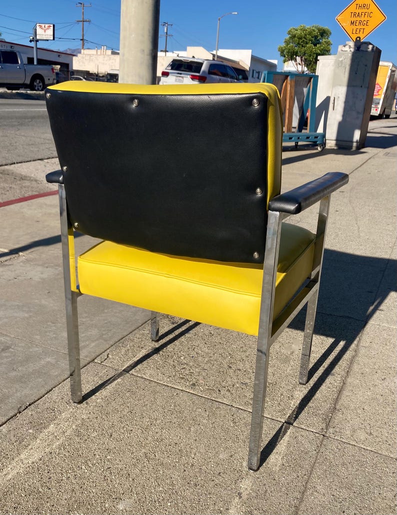 Op de afbeelding: Een retro-stoel met een heldergele zitting en rugleuning, gecontrasteerd door een zwarte rugleuning. De stoel heeft een verchroomd frame en armleuningen, wat een modern design uit het midden van de eeuw laat zien. De stoel staat buiten op een stoep.