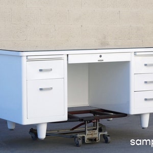 May include: White metal desk with a glass top and three drawers on each side. The desk is on a rolling dolly.