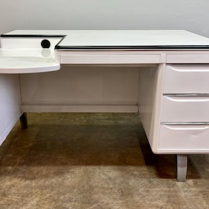 May include: A white desk with a black trim and three drawers. The desk has a curved side and a small shelf. The desk is made of wood and has silver legs. The desk is in a room with a neutral-colored wall.