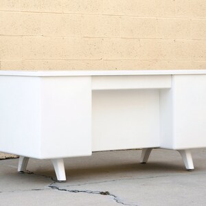 May include: A white metal desk with a simple design and four legs. The desk has two drawers on the left and right sides.