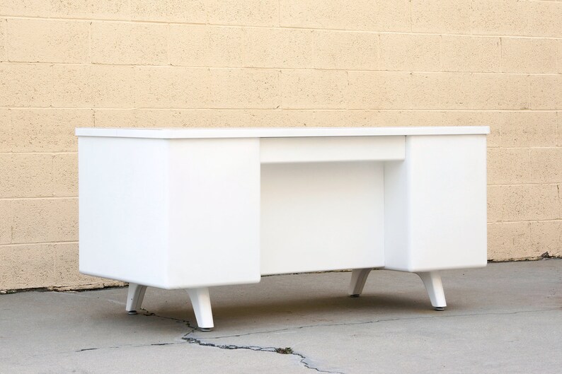 May include: A white metal desk with a simple design and four legs. The desk has two drawers on the left and right sides.