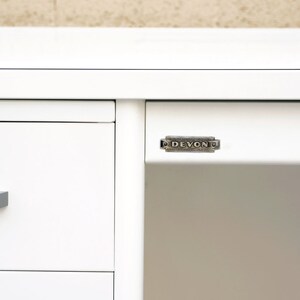 May include: White metal desk with three drawers and a silver metal nameplate that reads "DEVON" on the front right side.
