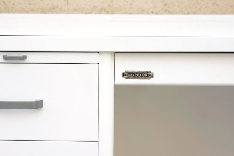 May include: White metal desk with three drawers and a silver metal nameplate that reads "DEVON" on the front right side.