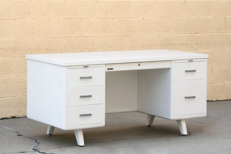 May include: White metal desk with three drawers on each side and a center drawer. The desk has a white laminate top and angled legs.