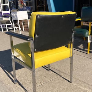 Op de afbeelding: Een retro stoel met een felgele zitting en een zwarte rugleuning. De stoel heeft een verchroomd frame en armleuningen. De rugleuning is rechthoekig en bevestigd met zilveren klinknagels. De stoel staat buiten voor een winkel.