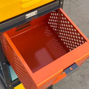 May include: A metal storage cabinet with a drawer that is open. The drawer is orange and has a perforated metal front. The cabinet is black and has yellow drawers above the orange drawer.