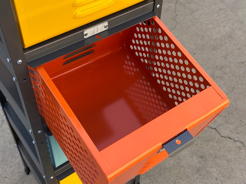 May include: A metal storage cabinet with a drawer that is open. The drawer is orange and has a perforated metal front. The cabinet is black and has yellow drawers above the orange drawer.