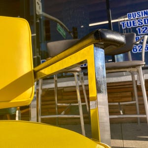 Op de afbeelding: Close-up van een gele en chromen kappersstoel. De stoel heeft een gele zitting en rugleuning met zwarte armleuningen. Het frame is chroom en geel. De achtergrond toont een winkelpui met tekst.