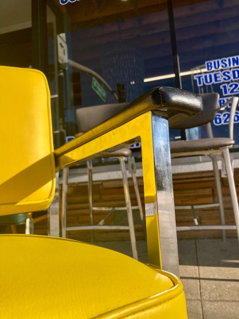 Op de afbeelding: Close-up van een gele en chromen kappersstoel. De stoel heeft een gele zitting en rugleuning met zwarte armleuningen. Het frame is chroom en geel. De achtergrond toont een winkelpui met tekst.