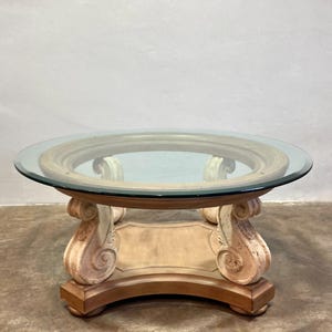 May include: Round coffee table with a clear glass top and a decorative base. The base features ornate, scroll-like supports in cream and brown tones. The table has a classic, elegant design, suitable for living rooms.