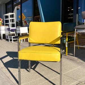 Op de afbeelding: Een gele gestoffeerde stoel met een chromen frame. De stoel heeft een rechthoekige zitting en rugleuning, met zwarte armleuningen. De stoel staat buiten, met andere stoelen zichtbaar op de achtergrond.