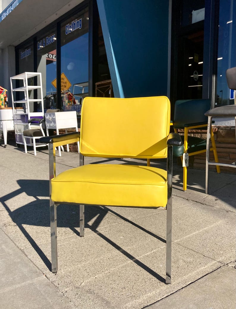 Op de afbeelding: Een gele gestoffeerde stoel met een chromen frame. De stoel heeft een rechthoekige zitting en rugleuning, met zwarte armleuningen. De stoel staat buiten, met andere stoelen zichtbaar op de achtergrond.