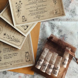 Puede incluir: Un conjunto de cinco frascos pulverizadores de vidrio transparente con etiquetas escritas a mano, contenidos en una bolsa de organza marrón con cordón, acompañados de varias tarjetas de papel crema con ilustraciones y texto en tinta negra, incluyendo Welcome to the Creamery Girls Correspondence Department.