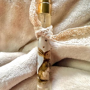 May include: A cylindrical glass perfume bottle with a gold-toned metal cap and a spray nozzle. The bottle features a label with a brown watercolor illustration of a rabbit and the partial text YED MOON. A cream-colored lace ribbon is tied around the neck.