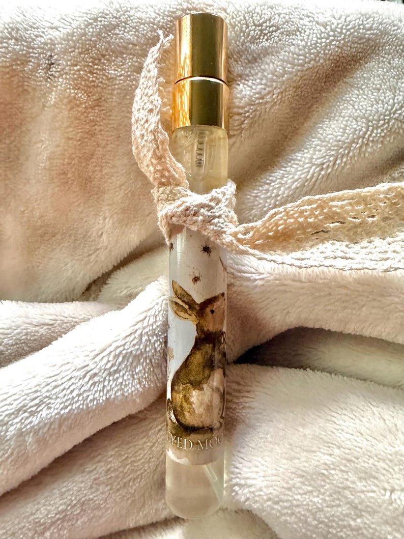 May include: A cylindrical glass perfume bottle with a gold-toned metal cap and a spray nozzle. The bottle features a label with a brown watercolor illustration of a rabbit and the partial text YED MOON. A cream-colored lace ribbon is tied around the neck.