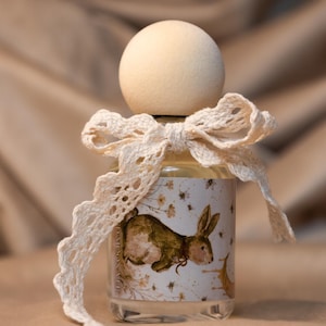 May include: A clear glass bottle with a round wooden cap and a cream-colored lace ribbon tied in a bow around the neck. The label features a brown rabbit illustration, small floral accents, and a crescent moon, with text reading D-MOON-VUE.