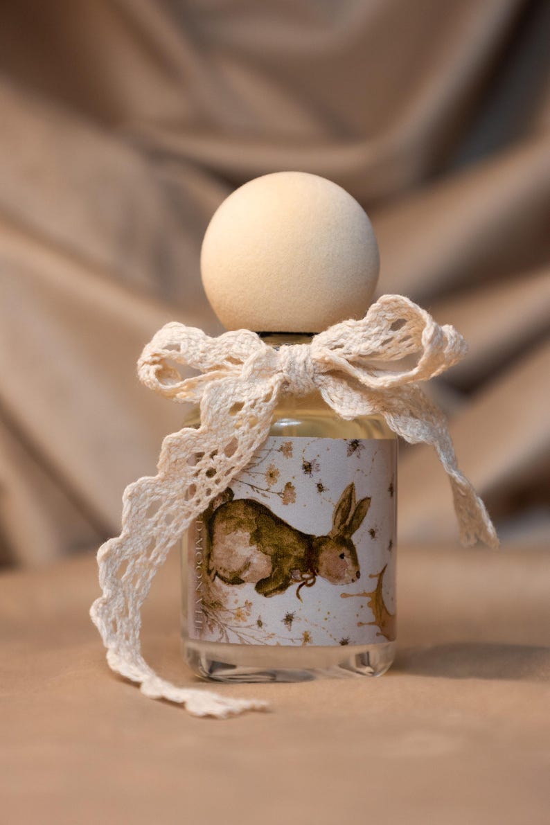 May include: A clear glass bottle with a round wooden cap and a cream-colored lace ribbon tied in a bow around the neck. The label features a brown rabbit illustration, small floral accents, and a crescent moon, with text reading D-MOON-VUE.