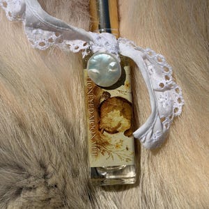 May include: A rectangular glass perfume bottle with a gold cap, featuring a label with the text HONEYED MOON VEIL and a botanical illustration. A white lace ribbon is tied around the neck, adorned with a single round, iridescent white pearl charm.