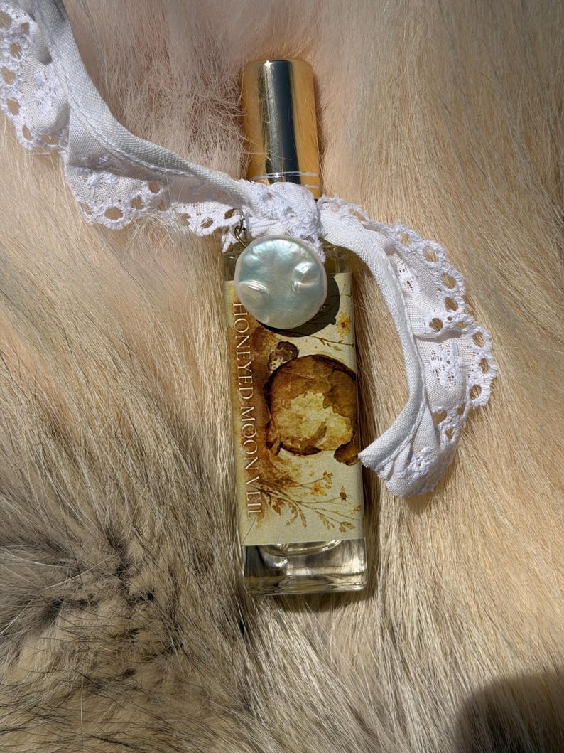 May include: A rectangular glass perfume bottle with a gold cap, featuring a label with the text HONEYED MOON VEIL and a botanical illustration. A white lace ribbon is tied around the neck, adorned with a single round, iridescent white pearl charm.