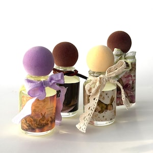 May include: Four glass perfume bottles with spherical caps in purple, brown, yellow, and brown. Each bottle features a unique label with rabbit illustrations and text, tied with decorative ribbons or lace bows around the neck of the glass container.