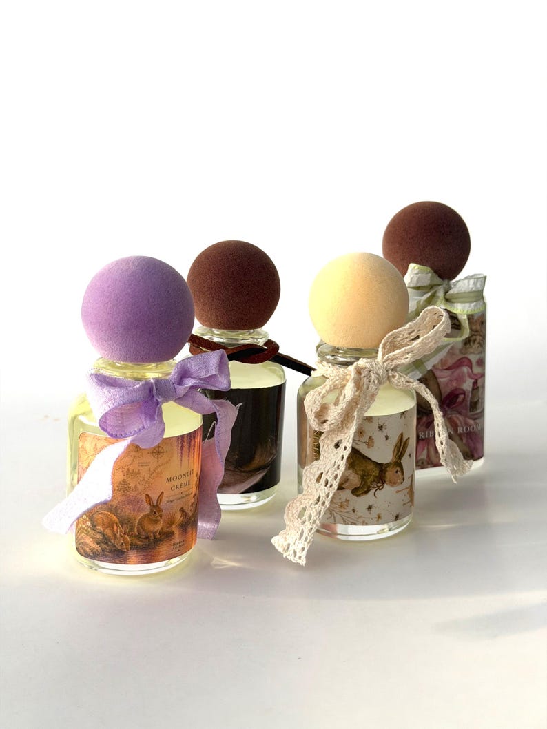May include: Four glass perfume bottles with spherical caps in purple, brown, yellow, and brown. Each bottle features a unique label with rabbit illustrations and text, tied with decorative ribbons or lace bows around the neck of the glass container.