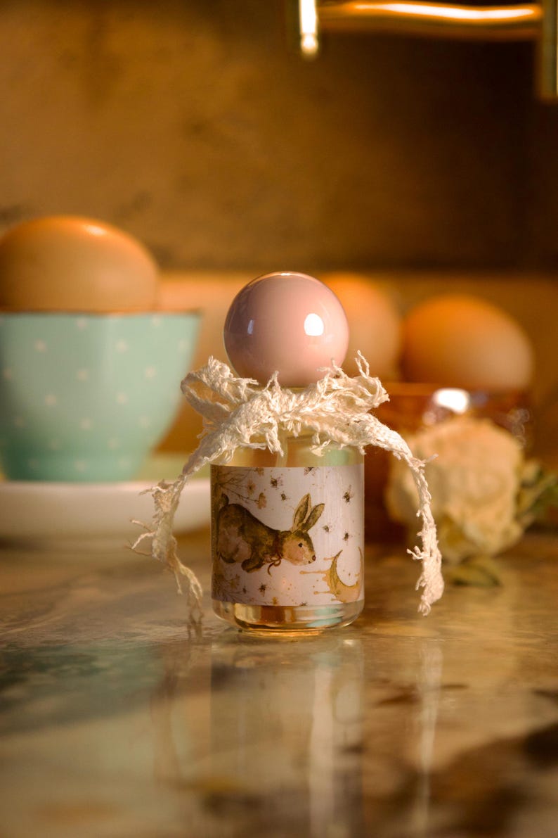 May include: A small clear glass bottle with a round light pink stopper. The bottle features a label with a brown rabbit illustration, small flowers, and a crescent moon. A frayed white fabric ribbon is tied around the neck of the bottle.