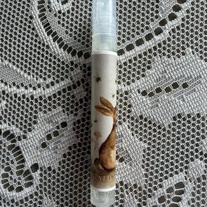 May include: A clear plastic spray bottle with a cylindrical shape and a transparent cap. The label features a watercolor illustration of a brown rabbit, small bees, and floral elements, with the text HONEYED MIST printed at the bottom.