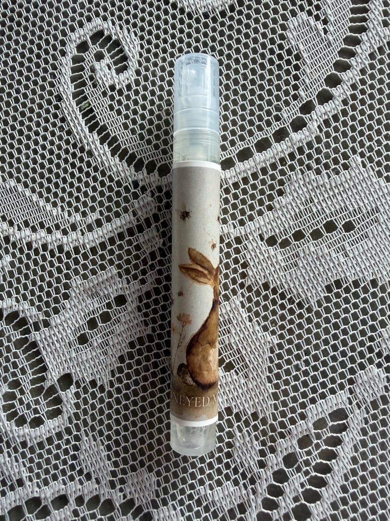 May include: A clear plastic spray bottle with a cylindrical shape and a transparent cap. The label features a watercolor illustration of a brown rabbit, small bees, and floral elements, with the text HONEYED MIST printed at the bottom.